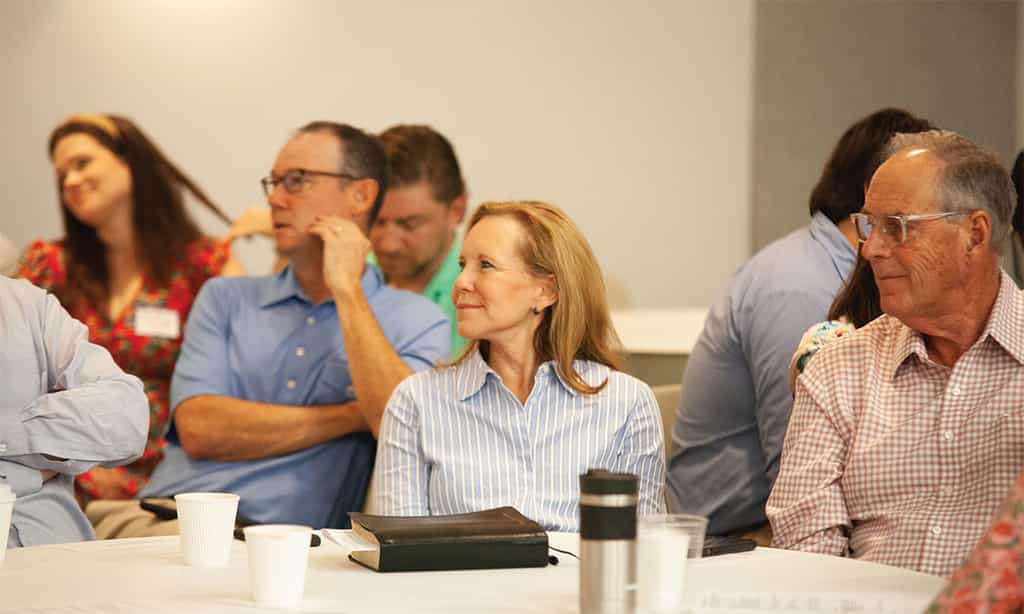 Adults seated at tables listening during a church class or discussion, with Bibles and coffee cups in front of them.