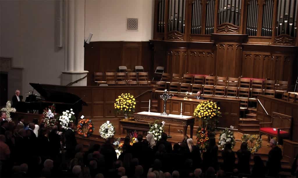 Congregation gathered in a church sanctuary for a memorial service, with a casket at the front surrounded by floral arrangements.