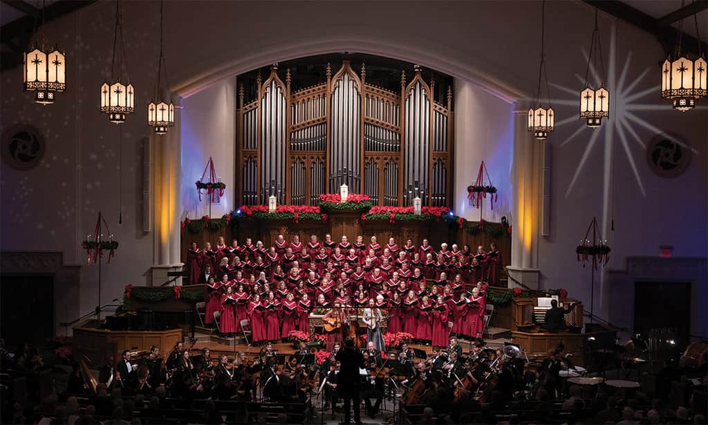 Large church choir in red robes singing in front of a pipe organ, accompanied by an orchestra.