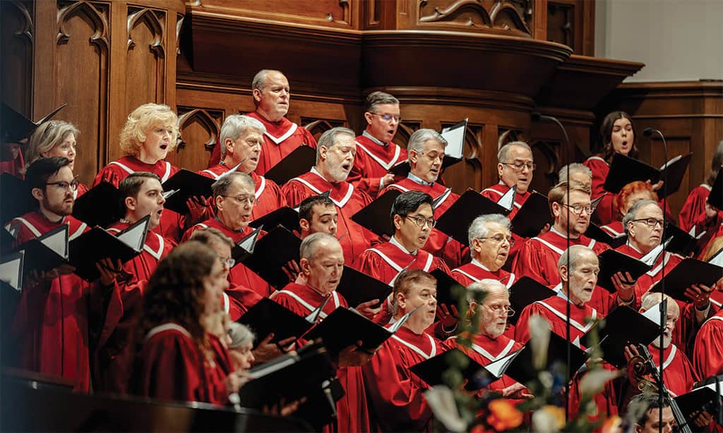Traditional church choir in red robes singing during a worship service in a wood-paneled sanctuary.