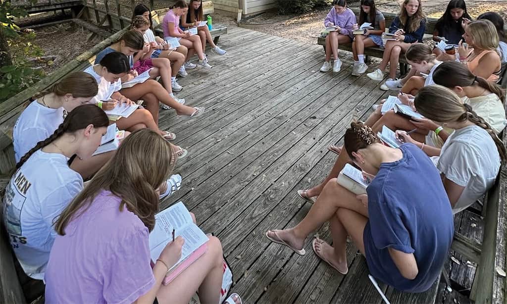 Teen girls gathered in a circle outdoors for Bible study, reading and writing in their books.