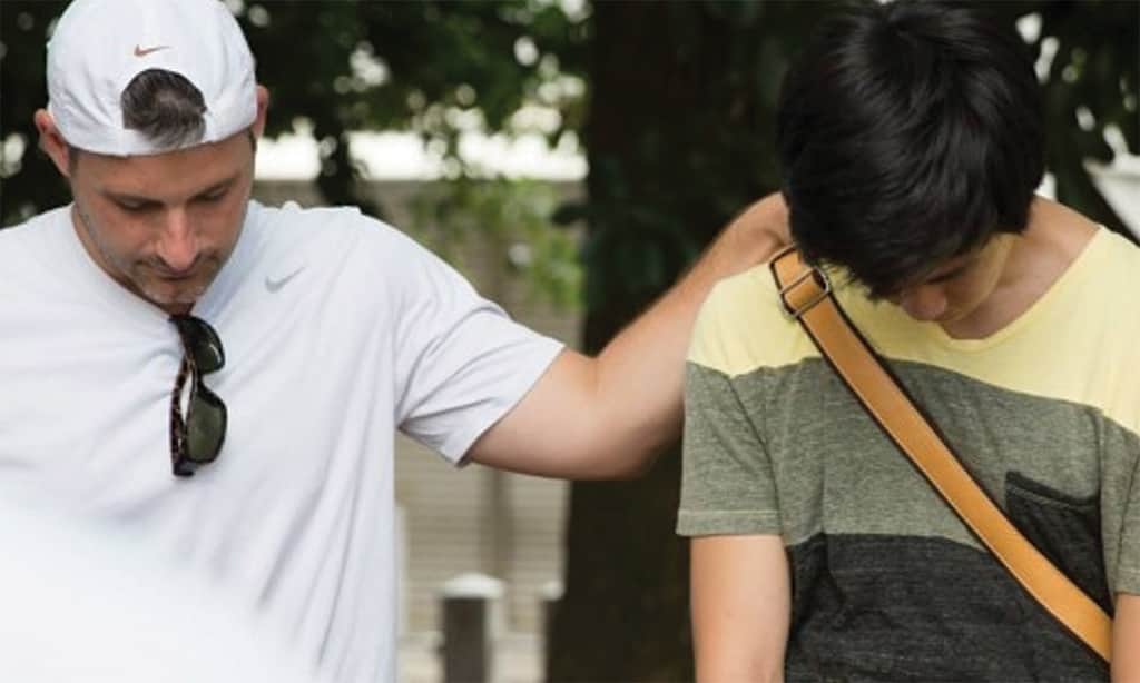 Man praying with teen outdoors, hand on shoulder.