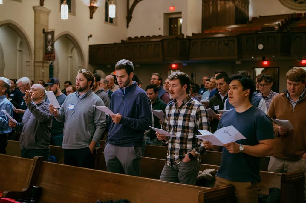 Men standing in church pews singing from printed programs during a worship service.