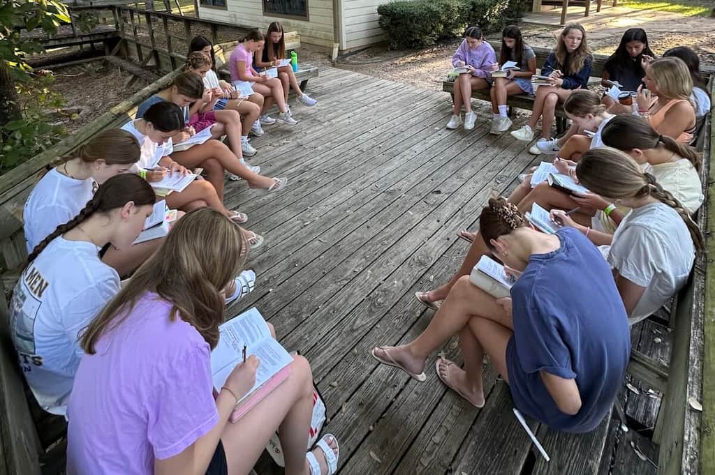Teen girls gathered in a circle outdoors for Bible study, reading and writing in their books.