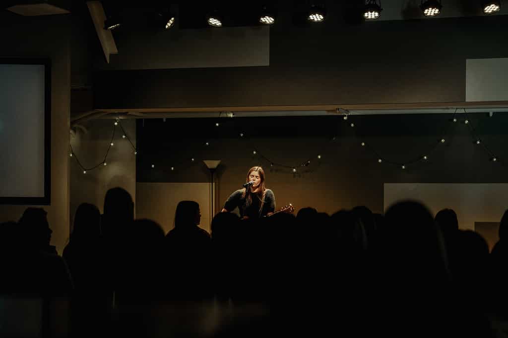 oung woman singing and playing guitar on stage under soft lights as students gather for a High School Ministry worship service.