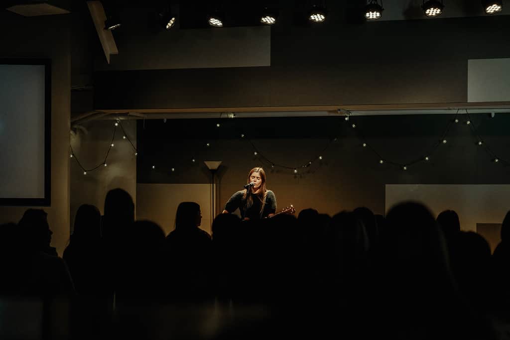 oung woman singing and playing guitar on stage under soft lights as students gather for a High School Ministry worship service.