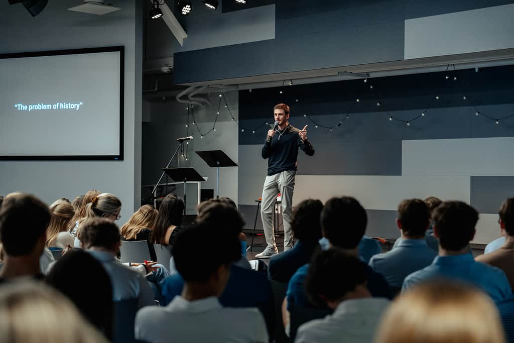 High school students seated and listening as a young man speaks with a microphone on stage at a Dallas High School Ministry.