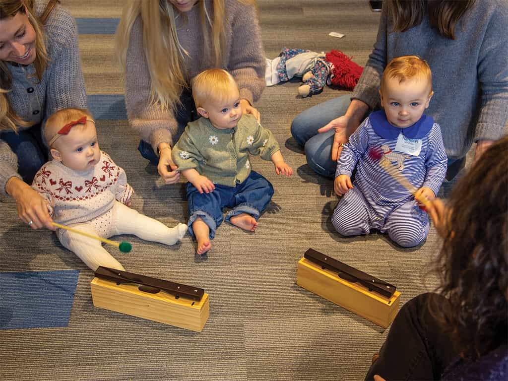 Babies sitting on the floor with caregivers playing simple instruments during music classes for babies in Dallas.