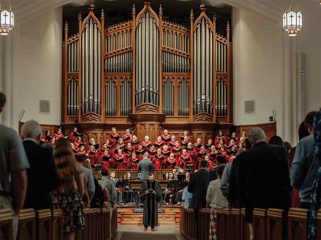 Church choir in red robes singing in the sanctuary beneath a large pipe organ, with the congregation standing during worship.
