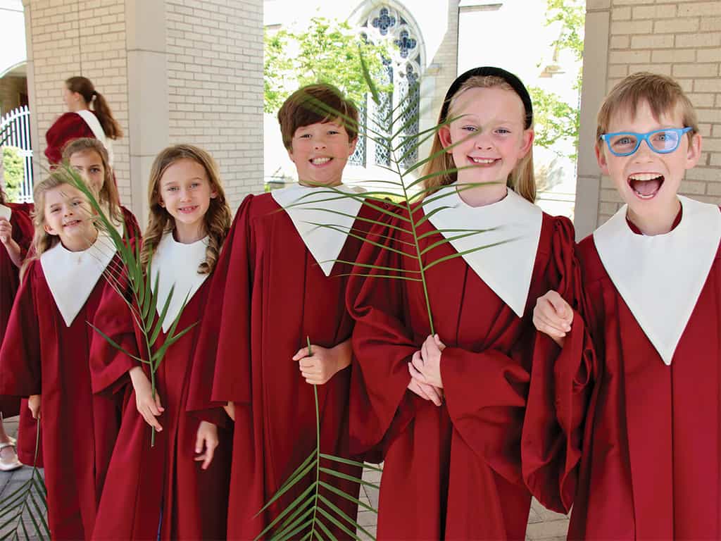Children in red choir robes holding palm branches outside a church, smiling during a Palm Sunday celebration.