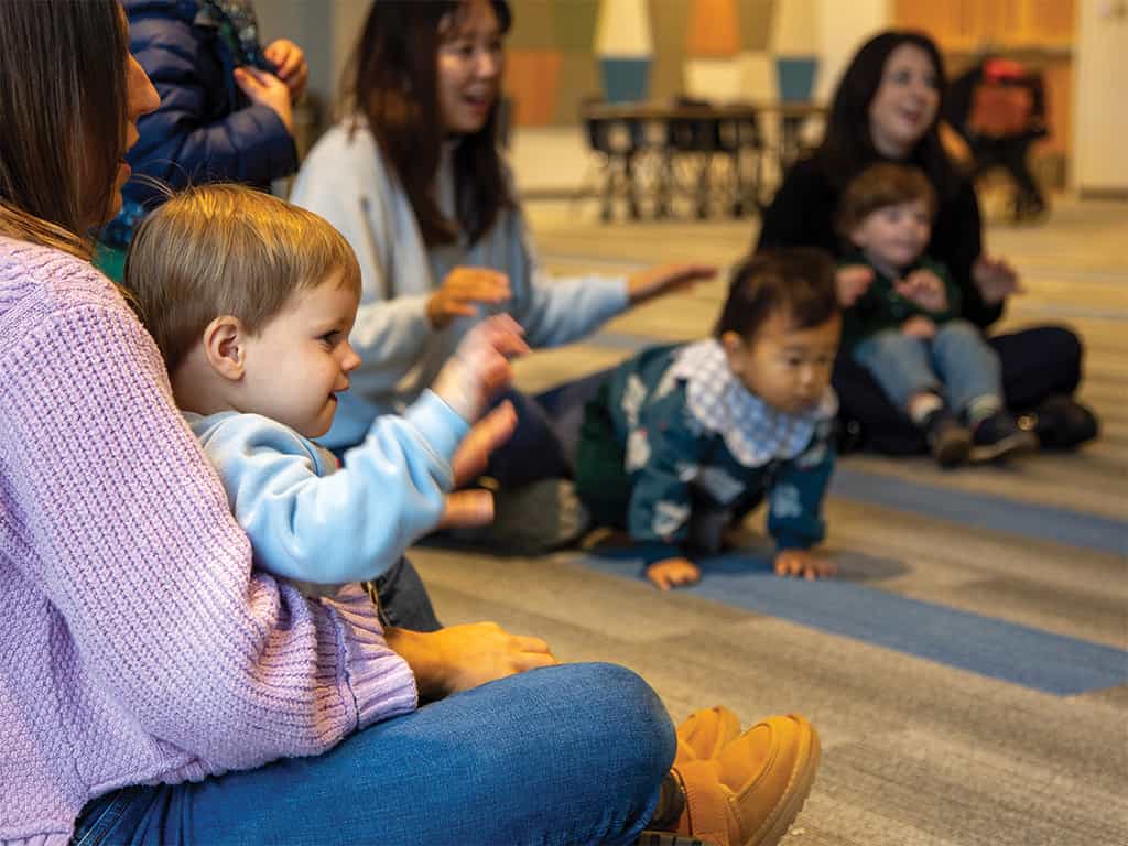 Toddlers sitting and crawling on the floor with caregivers during a parent-child group activity indoors.