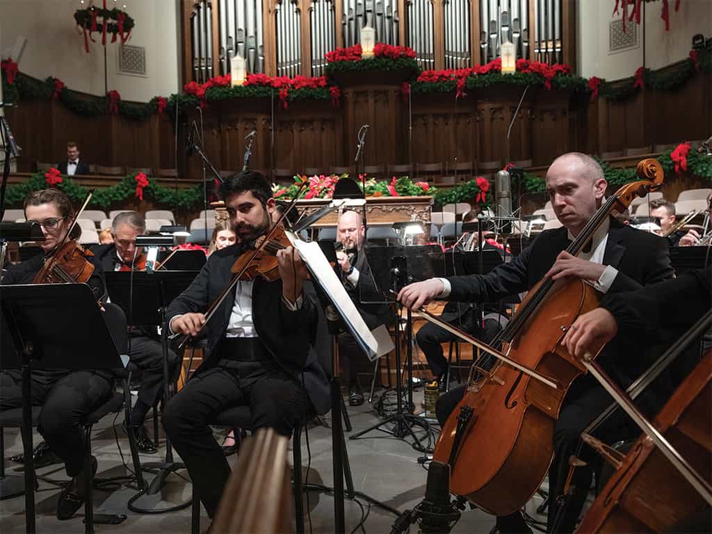 Orchestra performing a Christmas concert inside a church sanctuary, with musicians playing strings beneath festive red and green holiday decorations.