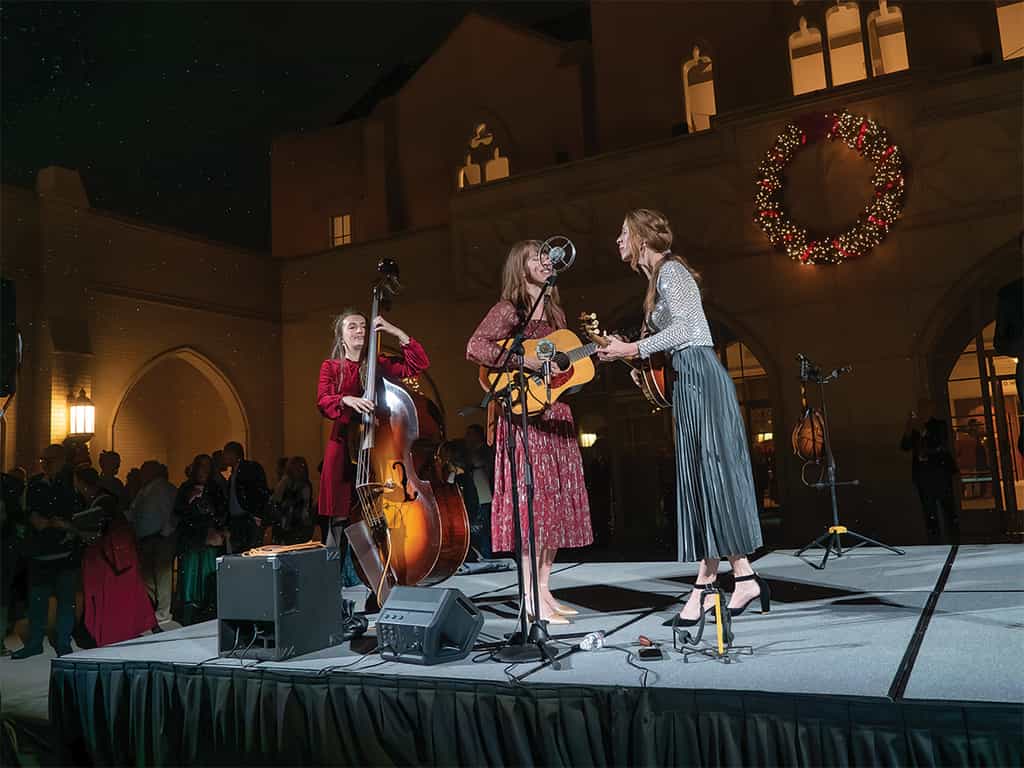 Live Christmas concert performance outside a church, featuring three musicians singing and playing guitar and upright bass beneath a large holiday wreath.