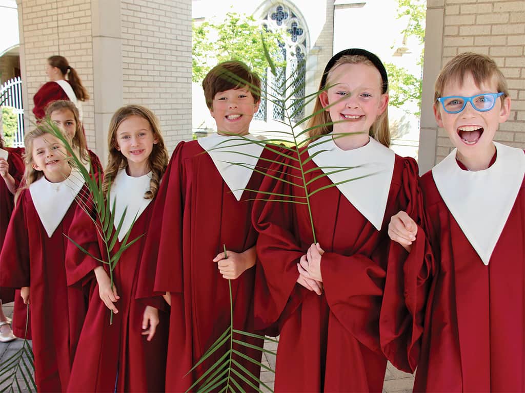 Children in red choir robes holding palm branches outside a church, smiling during a Palm Sunday celebration.