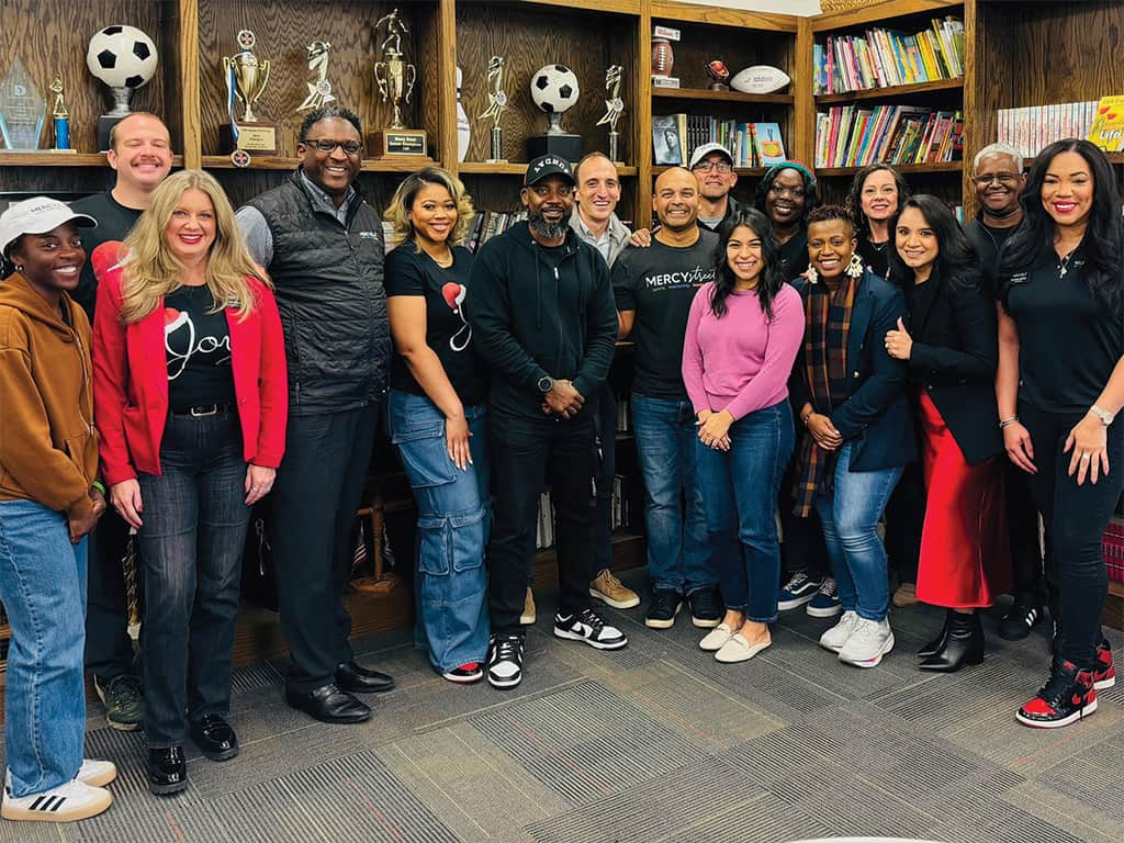 Diverse group of adults standing together and smiling in front of bookshelves filled with books and trophies.