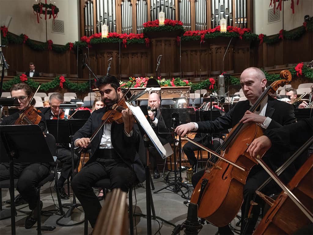 Orchestra performing a Christmas concert inside a church sanctuary, with musicians playing strings beneath festive red and green holiday decorations.