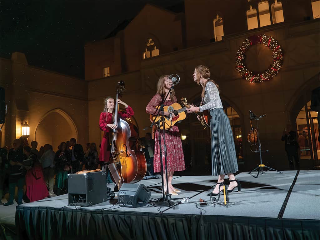 Live Christmas concert performance outside a church, featuring three musicians singing and playing guitar and upright bass beneath a large holiday wreath.