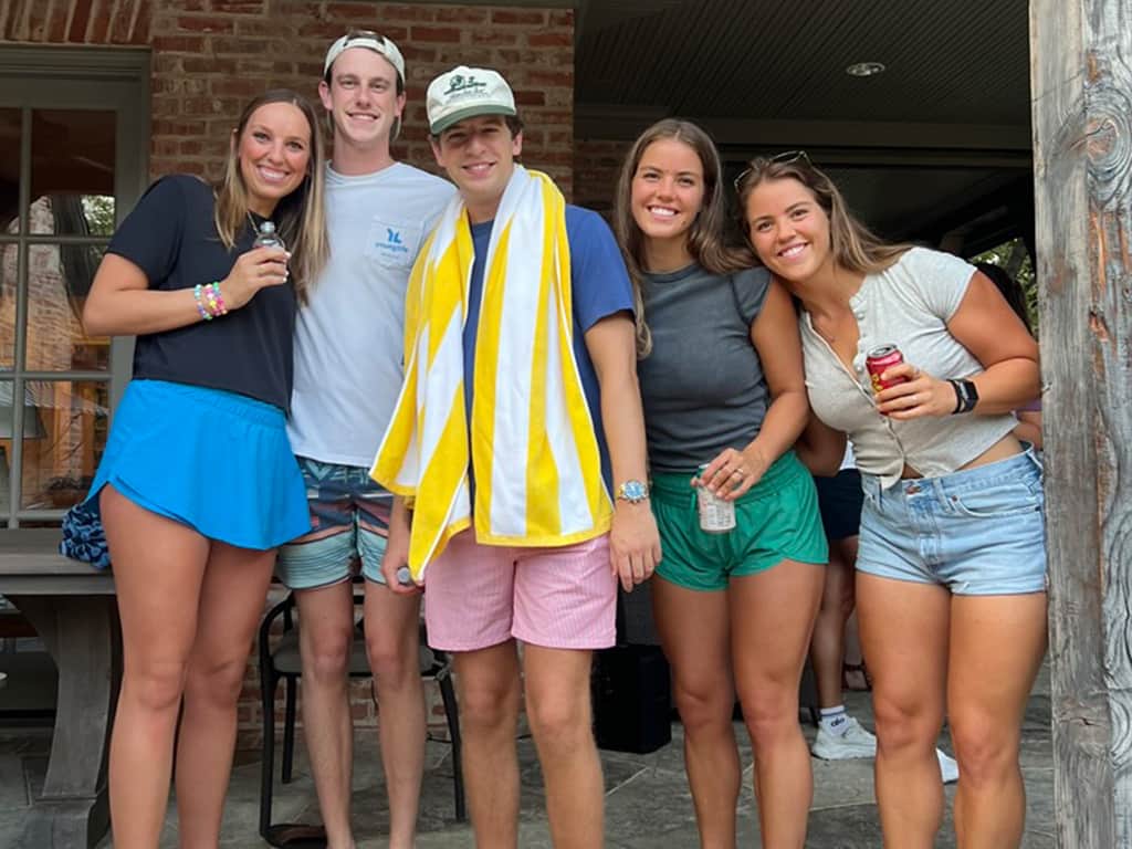 Five young adults wearing bright colored clothes and smiling together on a porch, enjoying time outdoors.