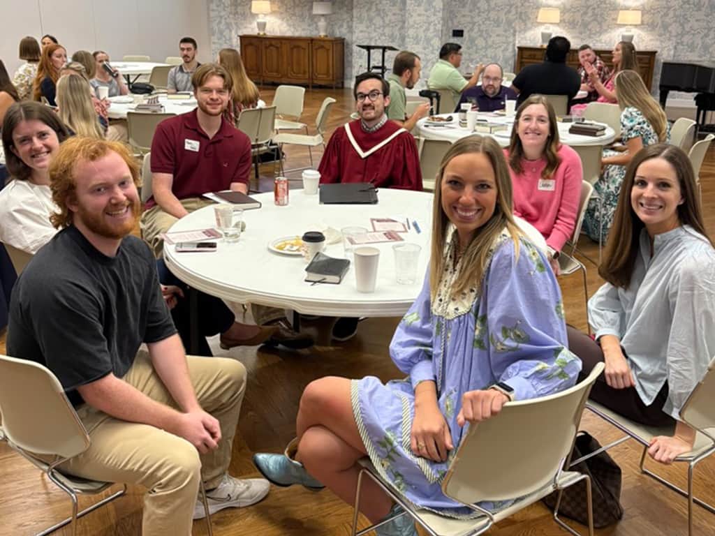 Small group of adults seated around a round table in a church fellowship hall, smiling during a Bible study or community gathering.