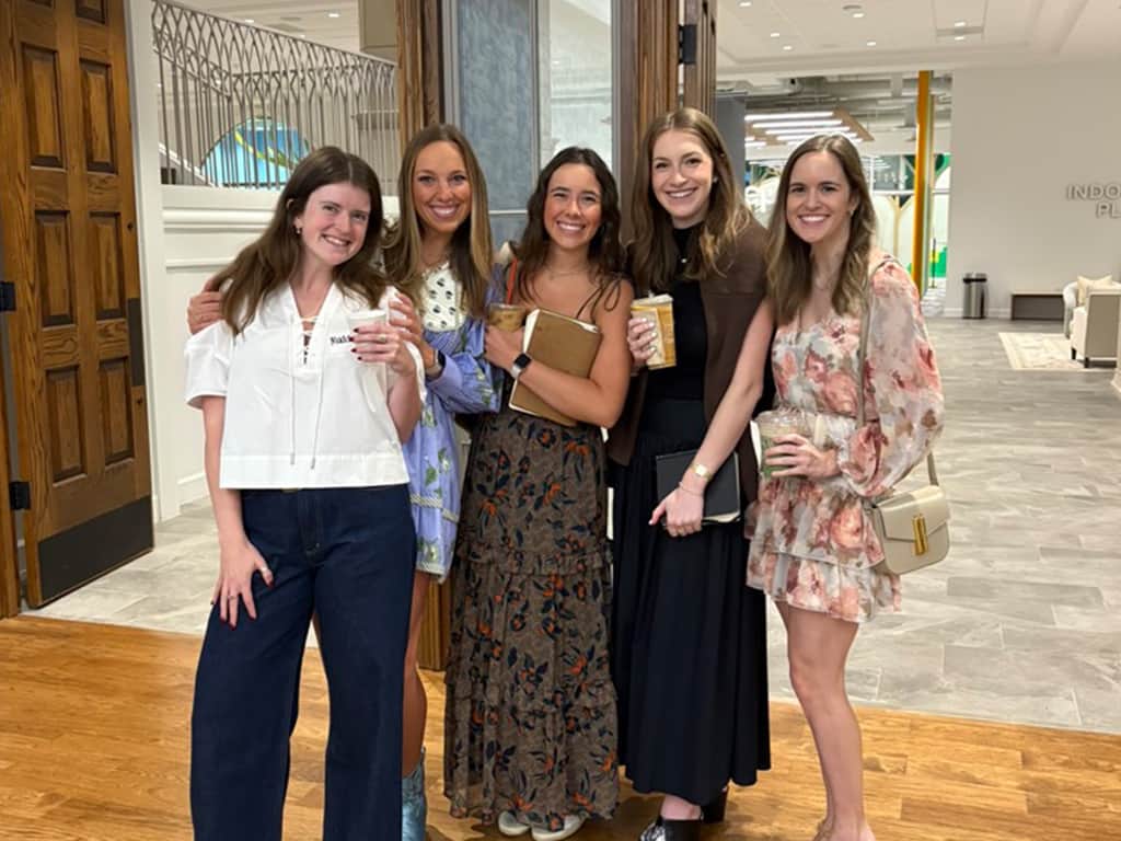 Five women standing together inside a church lobby, smiling and holding coffee and Bibles.