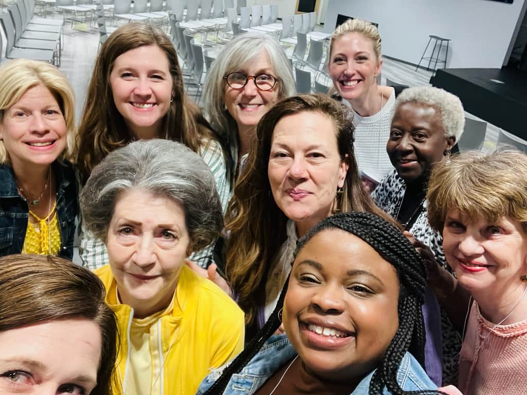 Group of women smiling together for a selfie in a church classroom with rows of chairs in the background.