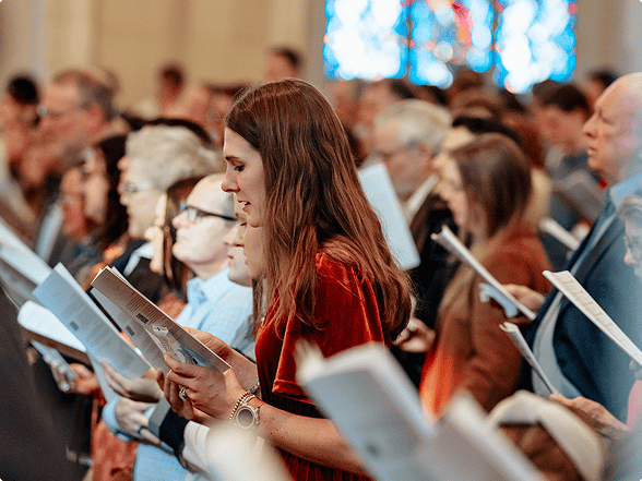 Congregation members reading from printed orders of worship during a church service, with stained glass windows in the background.