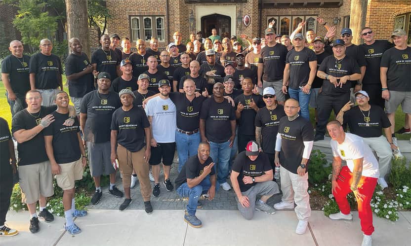 Large group of men wearing matching shirts standing together outside in front of a brick building.