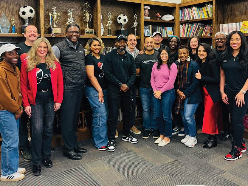 Diverse group of adults standing together and smiling in front of bookshelves filled with books and trophies.