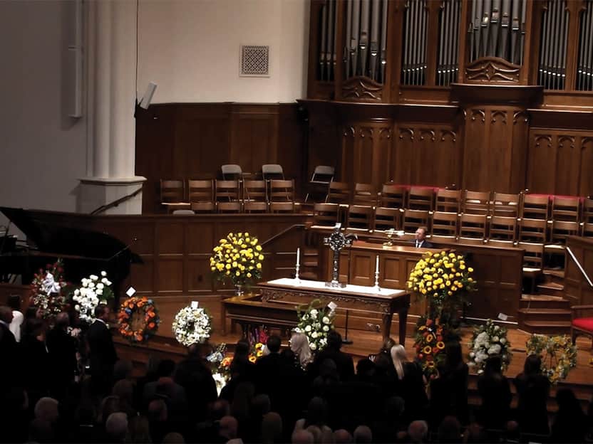 Congregation gathered in a church sanctuary for a memorial service, with a casket at the front surrounded by floral arrangements.